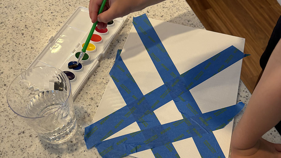 Child painting with watercolor paints in between the blue tape strips on white poster board.