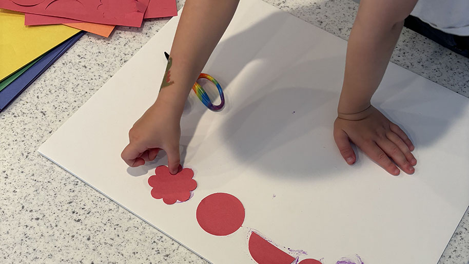Child placing colored construction paper shapes in lines on white poster board.