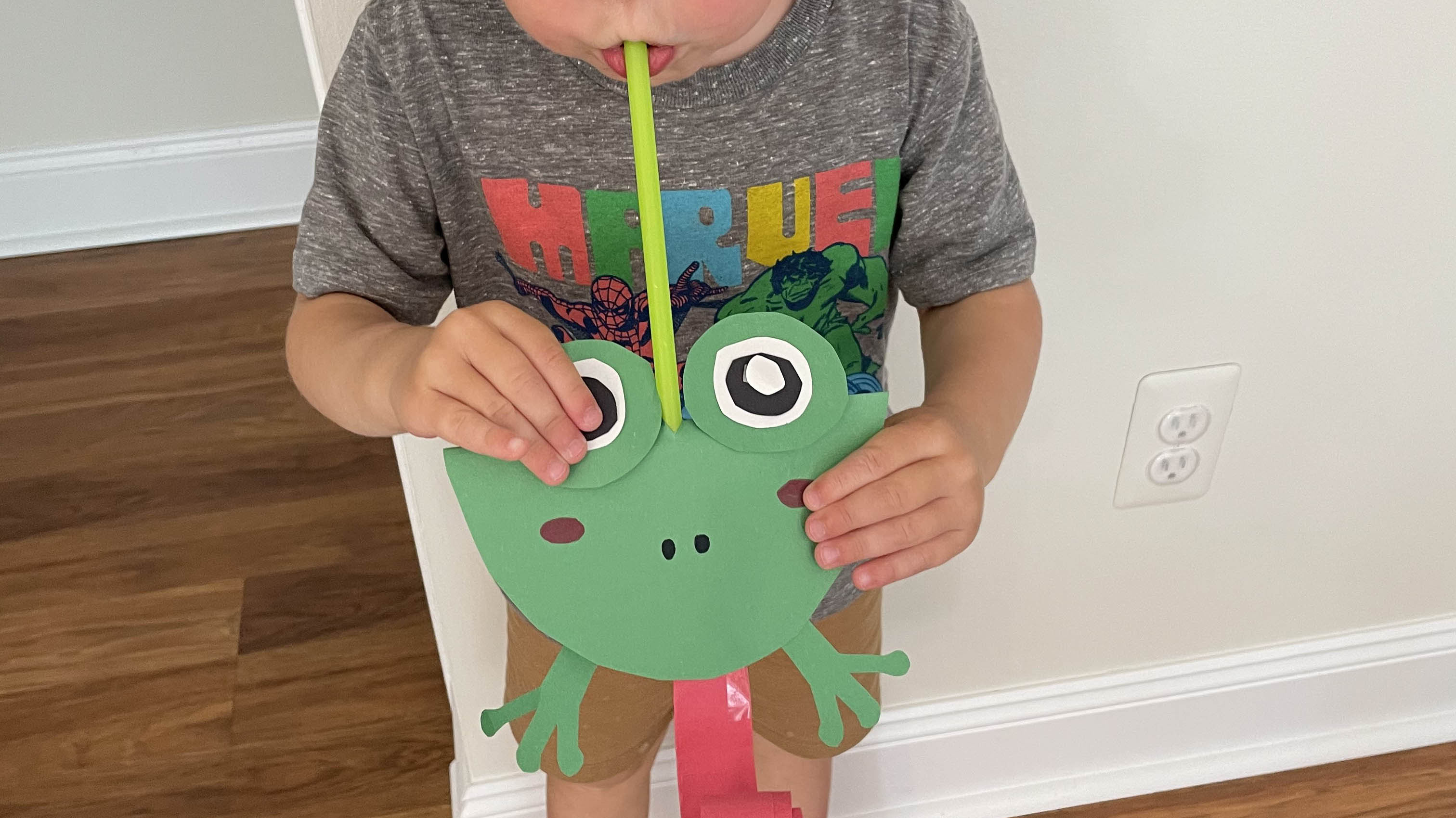 Child blowing into green straw of decorative paper frog to roll out red paper tongue