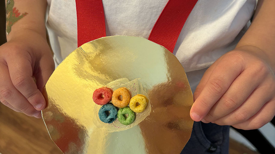 Child holding gold medal with red ribbon and cheerios of olympic ring design