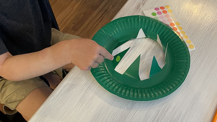 Adult helping child cut out every other triangle piece so that half of the triangles are outer portion of the paper plate and gap between each triangle.