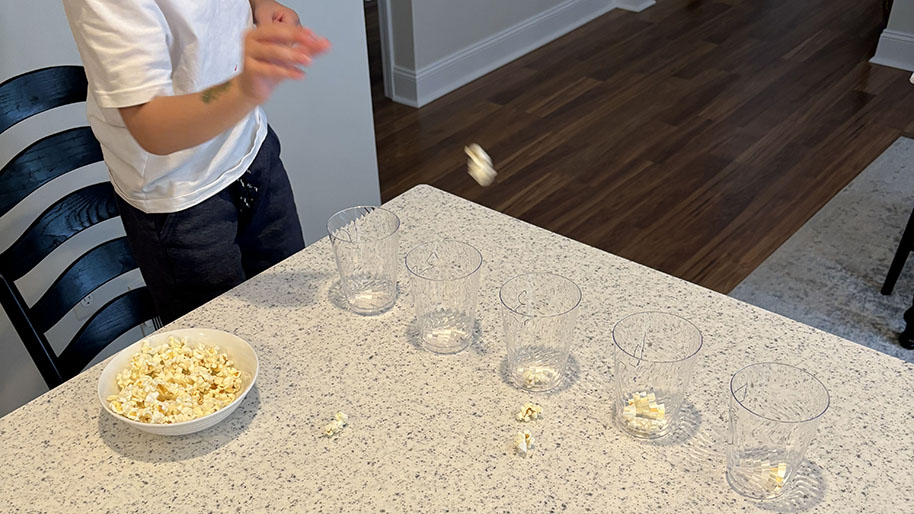 Child tossing popcorn into cups lined up on table.
