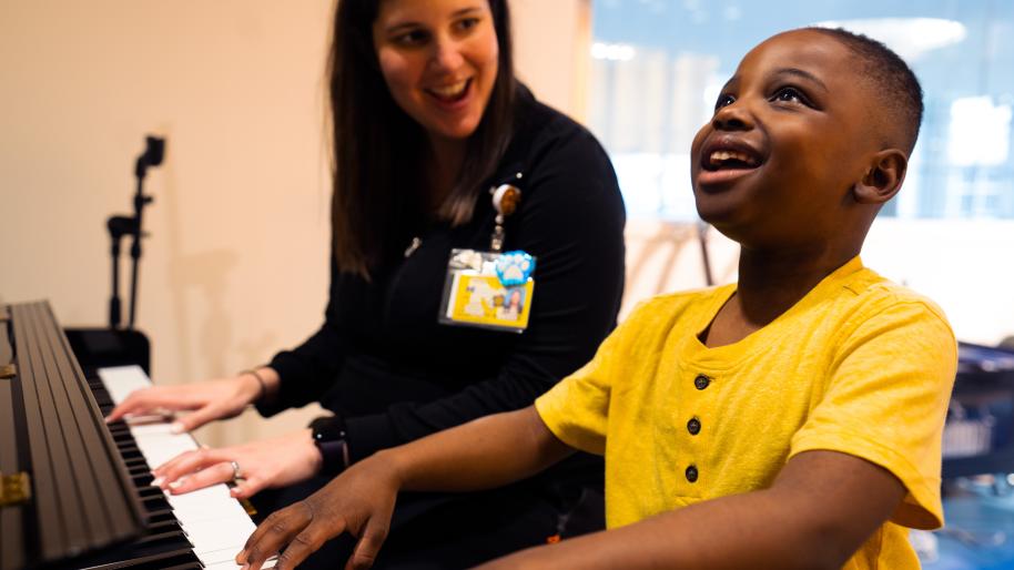 Young boy plays piano with female instructor smiling