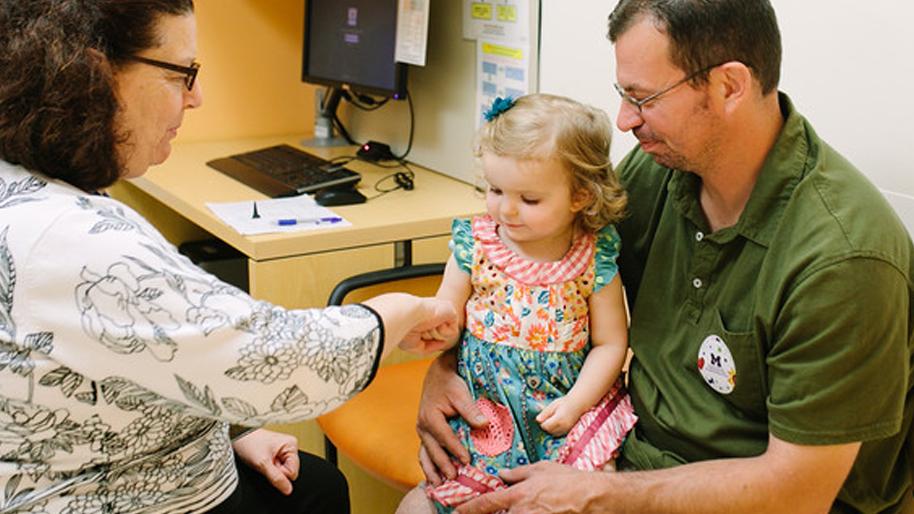 Little girl sitting on her father's lap while being examined by a doctor