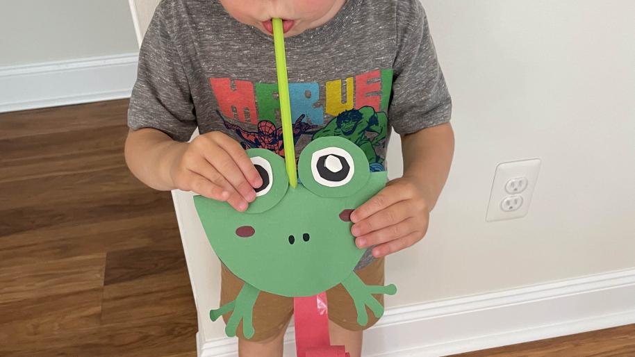 Child blowing into green straw of decorative paper frog to roll out red paper tongue