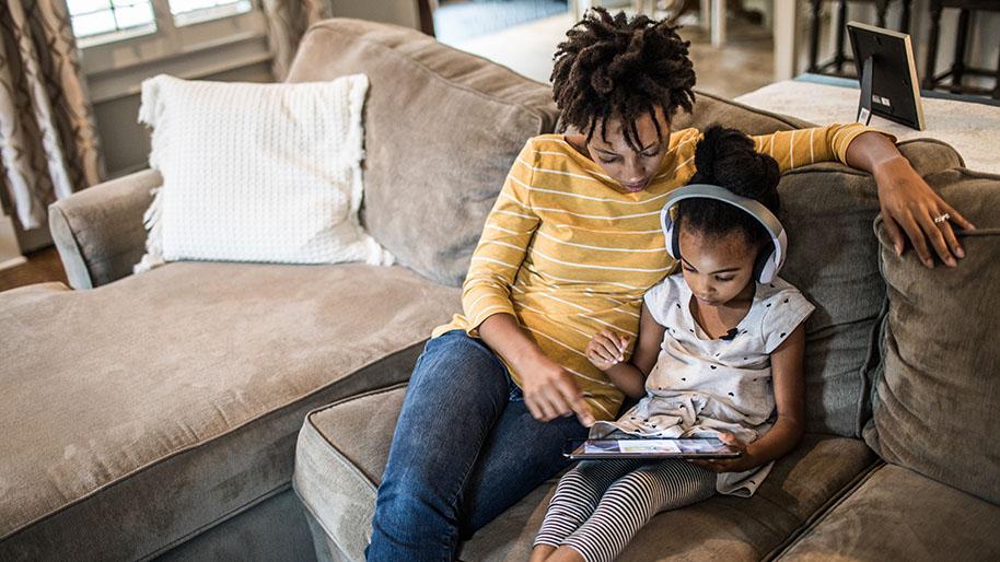 Mother and young girl holding an ipad sitting on a brown sofa.