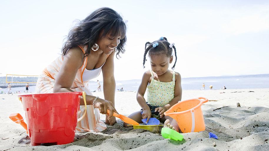 Mother and young girl playing in the sand with orange and yellow buckets; Ocean in the background