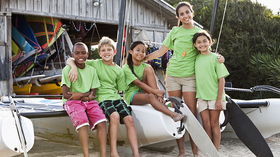 Two boys and two girls with there camp counselor sitting on a sailboat in the sand; boat house in the background